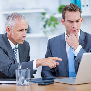 Two business men in an office environment where the older one is instructing the confused younger one by pointing at the laptop in front of them.