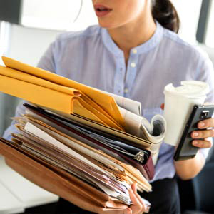 Extremely busy young business woman balancing too much paperwork and coffee cup in her hands.