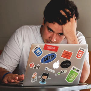 A young man stressed out and touching his forehead as he reads bad news on his laptop.